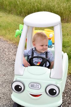 a little boy sitting in a toy car on top of gravel road with grass and bushes behind him