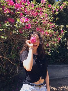 a woman standing in front of a bush holding a pink flower up to her face