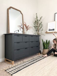 a woman is sitting on the floor in front of a dresser and mirror with plants