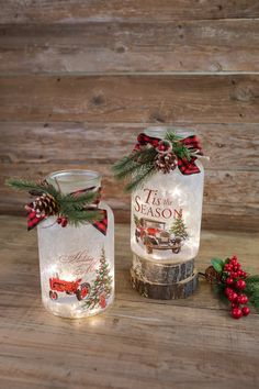 two glass jars with christmas decorations on them sitting on a wooden table next to holly and pine cones