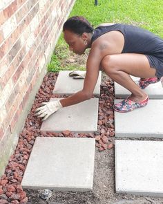 a woman kneeling down on cement blocks to lay bricks in the ground next to a brick wall