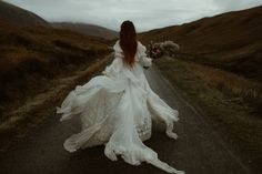 a woman is walking down the road wearing a long white dress and holding flowers in her hand