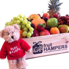 a teddy bear wearing a red sweater and standing next to a crate of fresh fruit