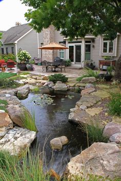 a small pond in the middle of a yard with rocks and grass around it, next to a house