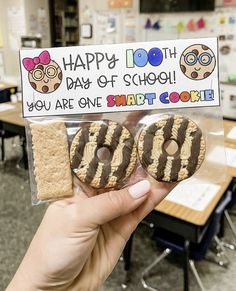 a person holding up two cookies in front of a sign that says happy 100th day of school you are give smart cookie