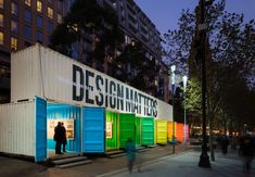 people are walking past colorful shipping containers on the sidewalk at night time, with buildings in the background