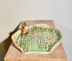 a small bird sitting on top of a green plate with trees in the background and a white wall behind it