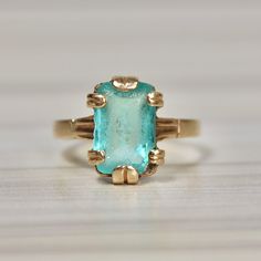 10K Yellow Gold, Vintage Retro Turquoise Ring, Synthetic Stone in bold setting, Size 8 Weight:  5.00 grams Ring Size:  8 Stone Measurements:  11.80 mm x 7.75 mm Shank:  2.55 mm This striking vintage ring, dating back to the 1930s or 1940s, features a captivating synthetic emerald cut stone set in a bold, retro-style setting. Its bright turquoise hue, reminiscent of old Hollywood glamour, is beautifully offset by the ring's chunky, statement-making design. Though the stone bears a small chip in the bottom right corner and light abrasion from age, these imperfections only add to its unique character, telling the story of its past. An estate piece with timeless charm, this ring effortlessly captures the essence of vintage elegance while making a daring, unforgettable statement. Classic Turquoise Ring For Collectors, Classic Turquoise Rings Collectible, Turquoise Emerald Ring For Anniversary, Collectible Art Deco Emerald Gemstone Ring, Vintage Yellow Gold Turquoise Ring For Wedding, Vintage Yellow Gold Turquoise Ring For Anniversary, Vintage Yellow Gold Turquoise Ring For Formal Occasions, Antique Turquoise Gemstone Ring For Formal Occasions, Vintage Turquoise Gemstone Ring For Formal Occasions