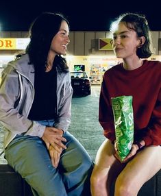 two women sitting next to each other in front of a store at night, one holding a bag of chips