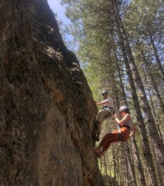 two people climbing up the side of a large rock in the middle of a forest