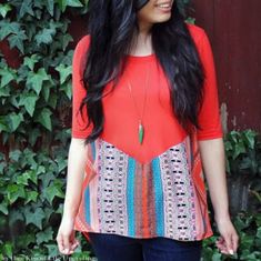 a woman with long black hair wearing a red shirt and jeans standing in front of a green bush