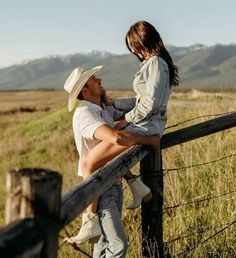 a man and woman leaning on a fence in an open field with mountains in the background