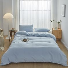 a bed with blue sheets and pillows in a white room next to a lamp on a table