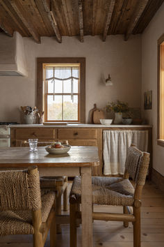 a wooden table sitting under a window next to a kitchen counter with two chairs and a bowl on top of it