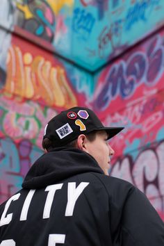 a man wearing a black hat with stickers on it standing in front of a graffiti covered wall