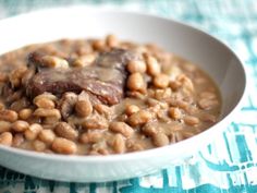 a white bowl filled with beans and meat on top of a blue table cloth next to a spoon