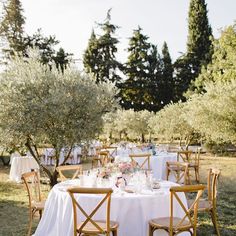 an outdoor table set up with white linens and wooden chairs, surrounded by trees