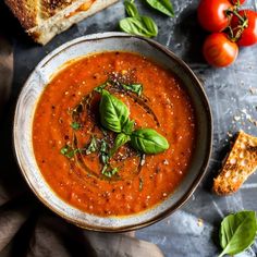 a bowl of tomato soup with bread and basil on the side, next to tomatoes