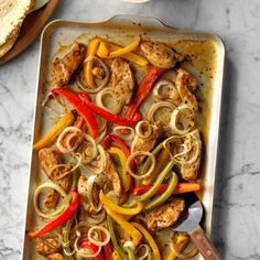 a pan filled with chicken and peppers on top of a marble counter next to bread