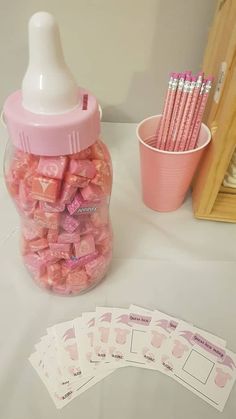 a jar filled with pink candy next to two cups and pencils on top of a table