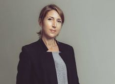 a woman standing in front of a gray wall wearing a black jacket and grey shirt