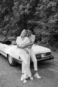 a man and woman sitting on the hood of a convertible car in front of trees