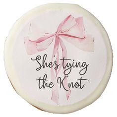 a round cake decorated with pink ribbon and saying she's tying the knot