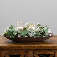 a wooden table topped with a bowl filled with white flowers and greenery next to candles