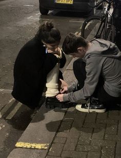 two people are sitting on the curb looking at their cell phones while one person stands over them