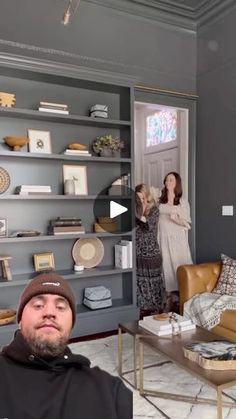 a man sitting in a living room next to a woman standing near a book shelf