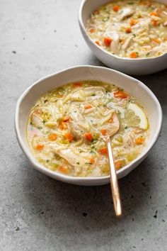 two bowls filled with chicken noodle soup on top of a gray counter next to a spoon