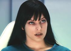 a woman with black hair and blue eyes is sitting in an office chair looking at the camera