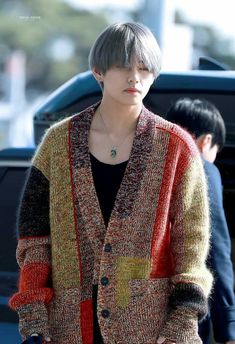 a woman with short hair wearing a multicolored cardigan and black shirt is standing in front of a car