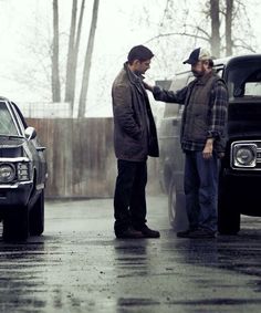 two men standing next to each other in front of parked cars on a rainy day