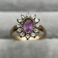 An adorable vintage amethyst and CZ cluster ring cast in 9-carat yellow gold. This gorgeous piece features a central oval-cut amethyst  in a beautiful shade of pink/light purple, encircled by 12 sparkling CZ stones. Such a charming, princess-worthy ring! Hallmarked by the London Assay Office, this ring has a maker's mark 'S&K' and bears a date stamp for 1989. It weighs 2.1 grams and fits a UK size L (US size 5.75). A complimentary ring box will be provided, making it ready for gifting or safekeeping. About Second Sparkle: Second Sparkle is my little collection of history & sparkle! As a jeweller's daughter with a historian's heart, I'm passionate about tales of times gone by and the sparkling legacy they left behind. Each piece in my store is a tiny moment from the past waiting to become p Oval Brilliant Cut Amethyst Ring In Yellow Gold, Classic Oval Amethyst Ring With Halo Setting, Antique Oval Cluster Ring With Halo Setting, Purple Cluster Rings For Formal Occasions, Formal Purple Cluster Rings, Vintage Purple Diamond Ring, Vintage Amethyst Ring With Diamond In Yellow Gold, Vintage Yellow Gold Amethyst Ring With Diamond, Vintage Oval Cluster Ring With Gemstone