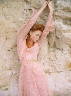 a woman in a pink dress leaning against a stone wall with her hands on her head