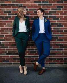 two people standing next to each other in front of a brick wall wearing blue suits