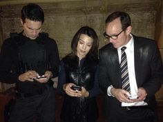 three people standing next to each other looking at their cell phones while one man in suit and tie looks down at his phone