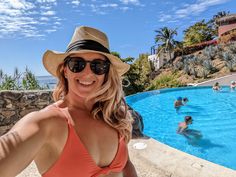 a woman taking a selfie in front of a pool with people swimming and sunbathing