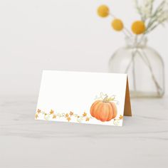 a white card with an orange pumpkin on it next to a vase filled with flowers