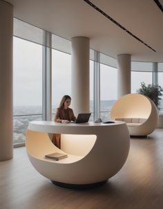 a woman sitting at a desk with a laptop computer in front of large windows looking out onto the city