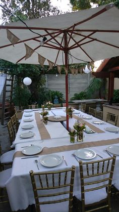 an outdoor dining table set up with white plates and place settings for six people under an umbrella