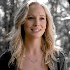 a woman with long blonde hair wearing a black jacket and smiling at the camera while standing in front of trees