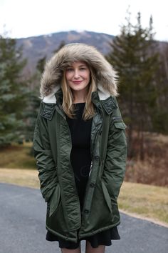 a woman standing in the middle of a road wearing a green parka and black dress