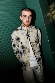 a man standing in front of a green wall wearing glasses and a tie dye shirt