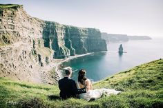 a man and woman sitting on top of a lush green hillside next to the ocean