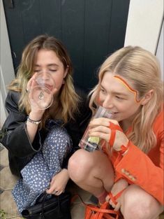 two young women sitting on the ground drinking from wine glasses and looking at each other