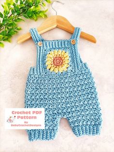 a crocheted blue overall with a yellow flower on the front and bottom, sitting next to a wooden hanger