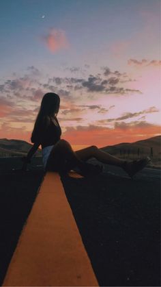 a woman sitting on the edge of a road at sunset with her legs spread out