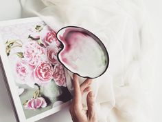 a hand holding a bowl with flowers on it next to an open photo book and white sheets